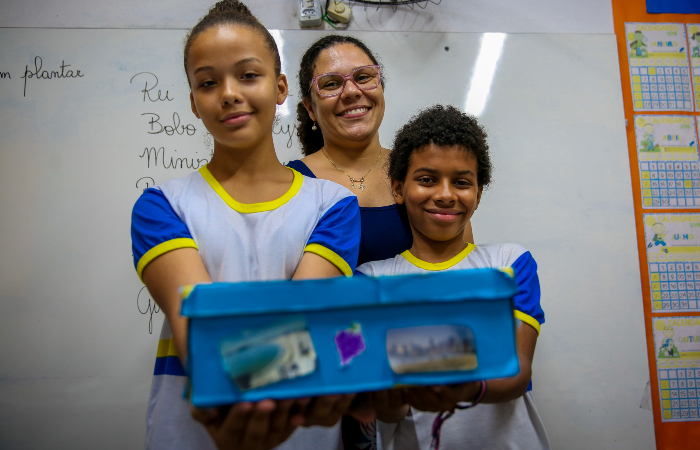 Guilherme junto com a colega Anna Beatryz, a professora Pâmela e o trabalho de geografia nota 10.