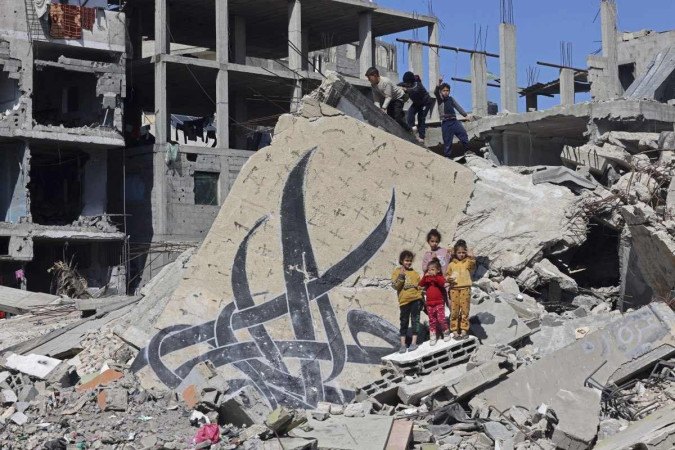 Crianças posam ao lado de um mural que restou em escombros de um edifício destruído em Rafah/crédito: MOHAMMED ABED / AFP