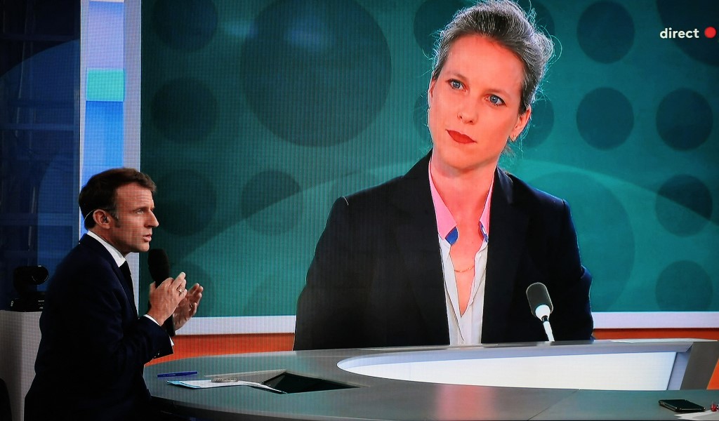 Presidente da França, Emmanuel Macron, dando uma entrevista ao vivo enquanto um retrato de Lucie Castets é exibido em uma tela gigante/Foto: LUDOVIC MARIN / AFP