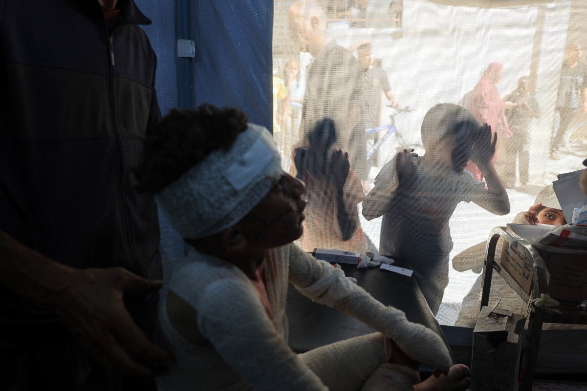 Crian&ccedil;a espia uma &aacute;rea de tratamento enquanto um palestino ferido est&aacute; sentado no Hospital al-Awda, em julho de 2024/Foto: Eyad BABA/AFP